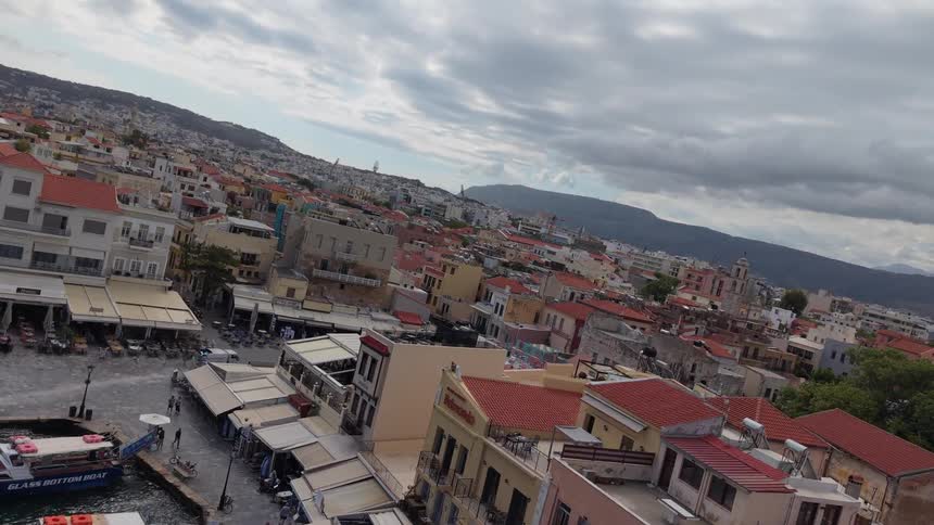A slow-motion aerial flyover showcases the charming waterfront and densely packed buildings of a historic Greek coastal town under a cloudy sky