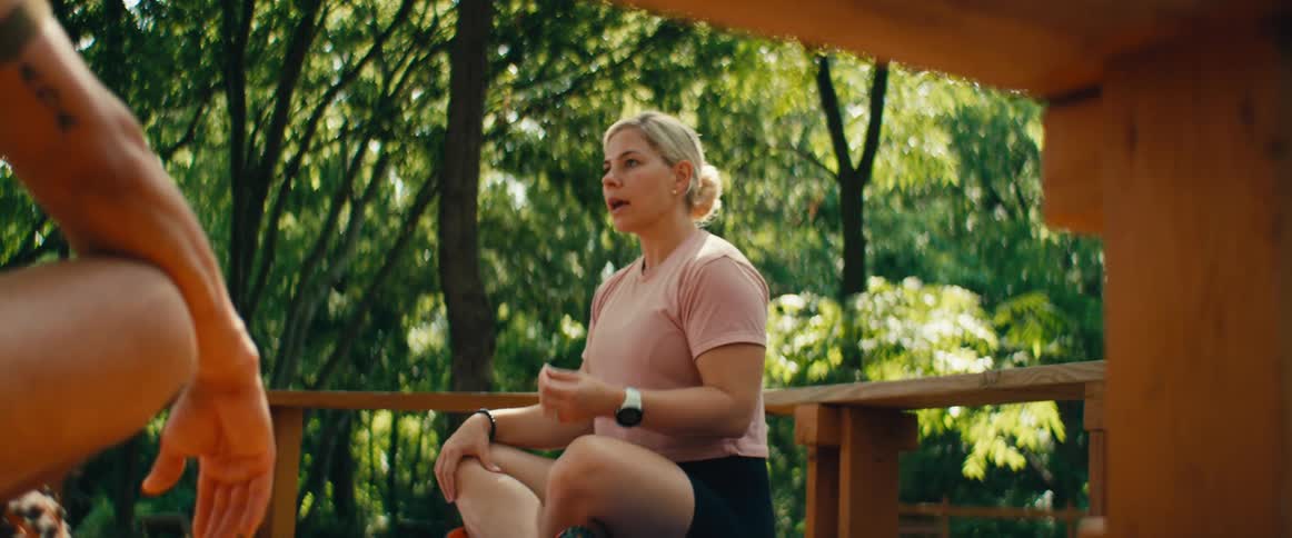 A woman in athletic wear leads a guided meditation and breathing exercise on a wooden deck surrounded by lush green trees