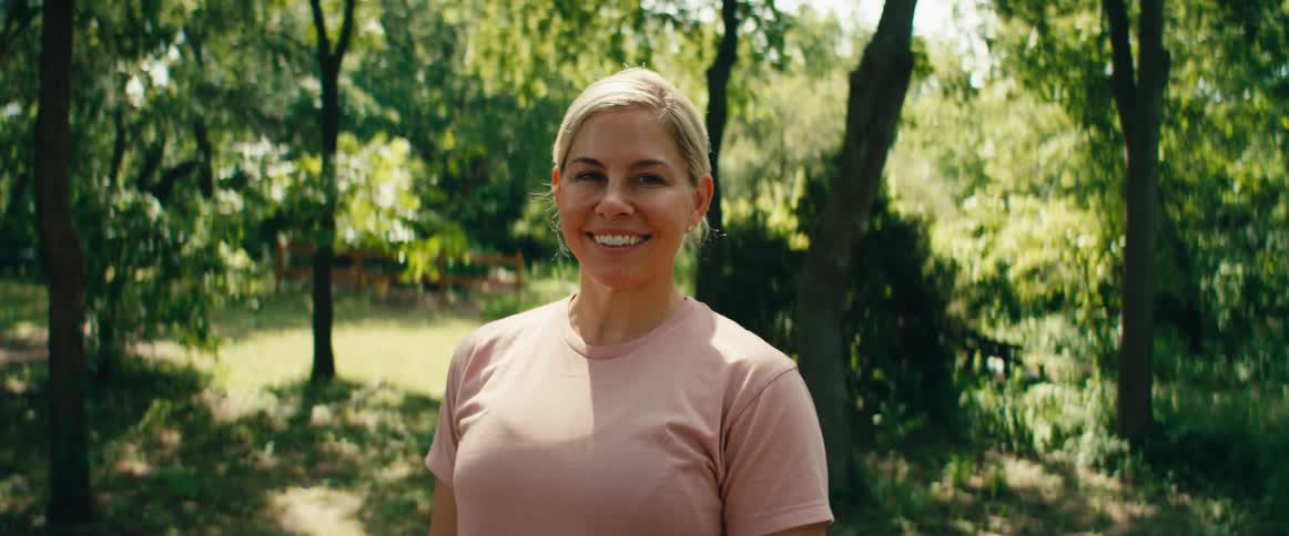A medium shot portrait of a happy blonde woman smiling while standing in a sunny, green park
