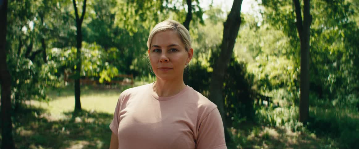 Portrait of a blonde woman in a pink shirt looking at the camera while standing in a sunlit park