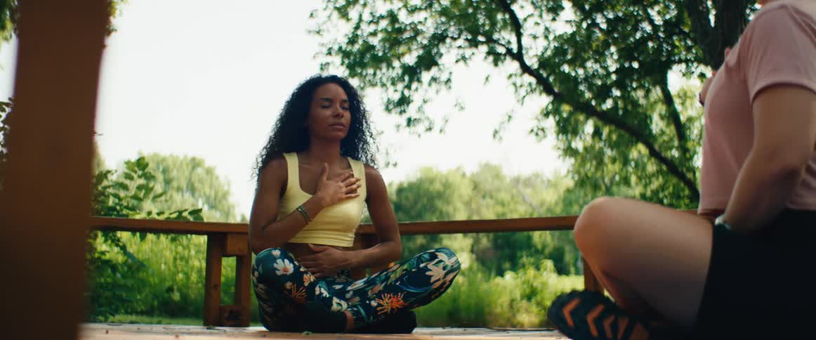 A peaceful woman with curly hair practices mindful breathing meditation while sitting on a wooden deck in nature