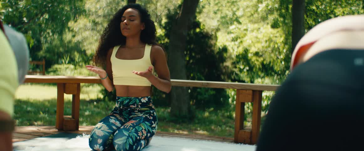 A focused woman with flowing curly hair practices yoga poses before transitioning into a calm meditation on a wooden deck in a sunlit park