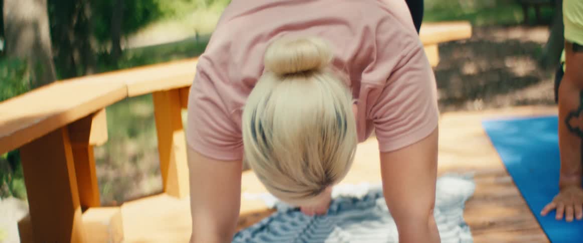 A blonde woman in a bun holds a downward dog yoga pose on a striped towel while stretching outdoors on a wooden deck