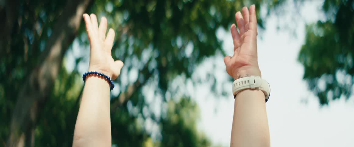 A serene blonde woman raises her arms and brings her hands into a prayer pose while meditating in a sunny park