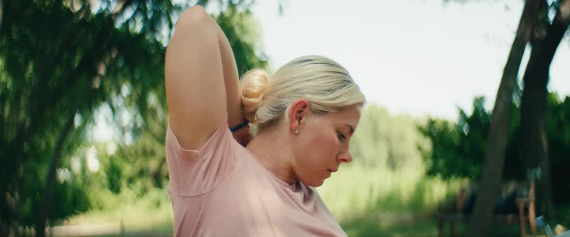 A close-up profile shot of a focused blonde woman stretching her arms and shoulders while sitting on a park bench