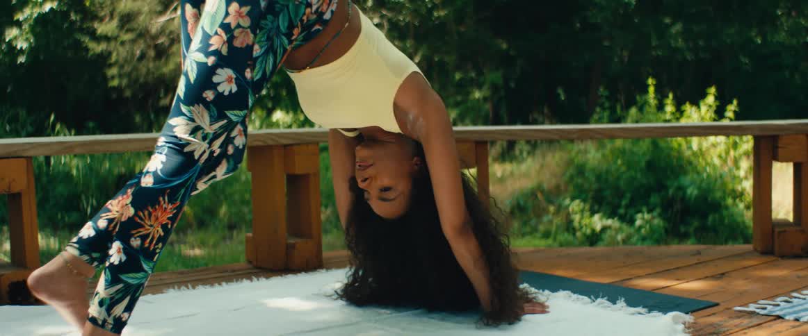 A smiling woman with long curly hair gracefully performs a downward-facing dog yoga pose in slow motion on a wooden deck outdoors