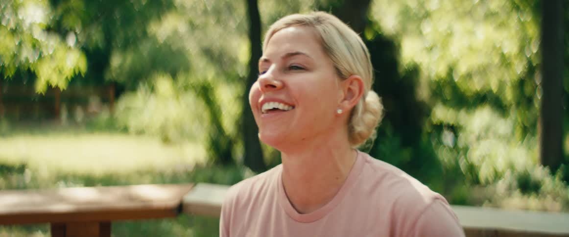A blonde woman in a pink shirt has a conversation outdoors, her expressions shifting from happy to serious