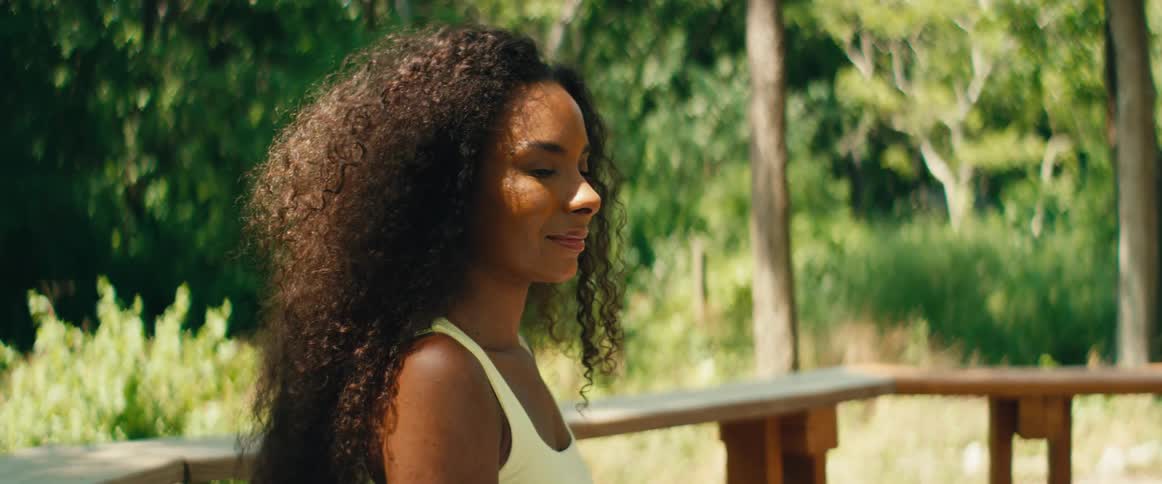 A serene woman with curly hair meditates outdoors, smiling as she closes her eyes and enjoys the warm sunlight
