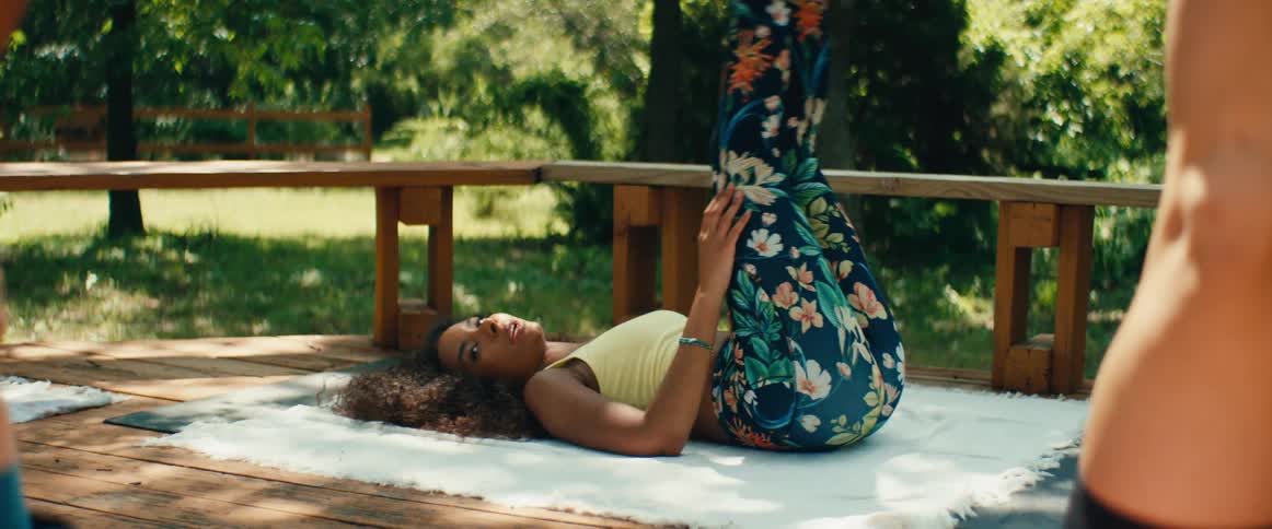 A young woman practices a supported shoulder stand yoga pose on a wooden deck in a lush, green outdoor setting