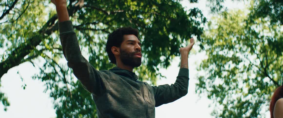 A young man in a hoodie raises his arms in meditation before settling into a tree pose on a yoga mat outdoors