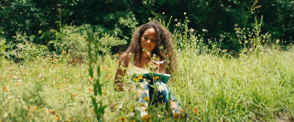 A creative young woman sits in a sunny meadow full of wildflowers and peacefully journals her thoughts