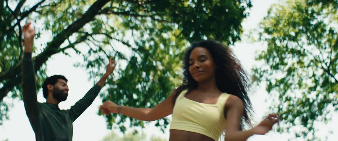 A young woman with beautiful curly hair smiles and dances expressively in a sunlit park, joined by her friends in a slow-motion sequence