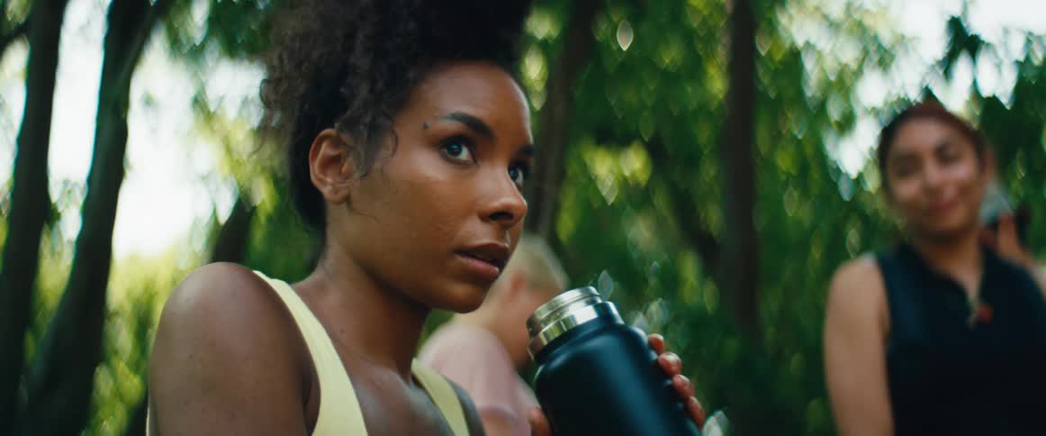 A thoughtful young woman with curly hair enjoys a quiet moment and drinks from her water bottle in a lush, sunlit park