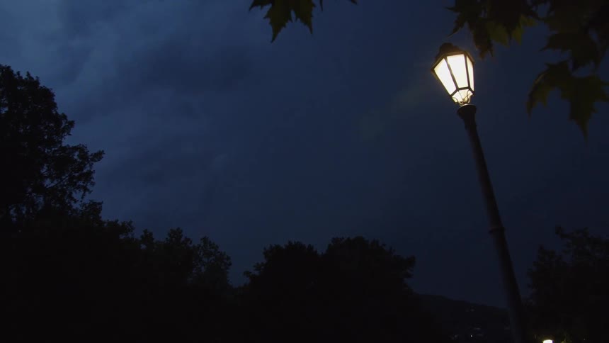 A single glowing lamppost stands against a dark and stormy night sky as lightning flashes dramatically in the clouds