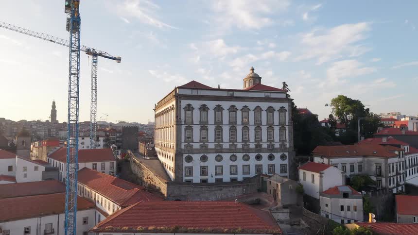 Aerial orbit of the historic Episcopal Palace in Porto, Portugal, at sunrise, with construction cranes and city rooftops in the background