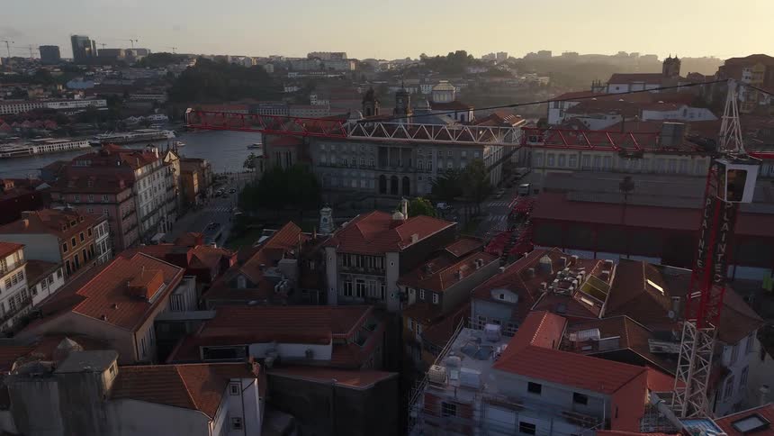 Pushing forward over the beautiful red rooftops of Porto, Portugal, as a crane stands tall over the city with the Douro River in the distance at sunset