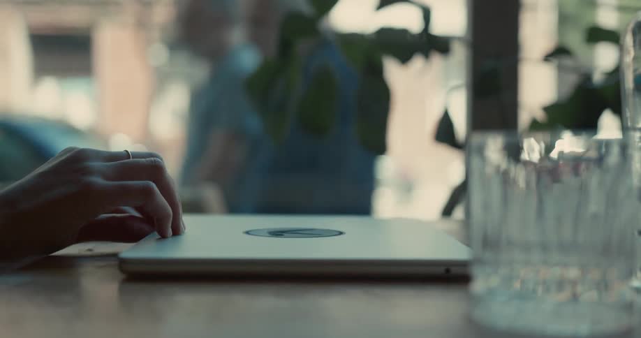 A close-up shot of a hand closing a silver laptop on a wooden table in a brightly lit cafe