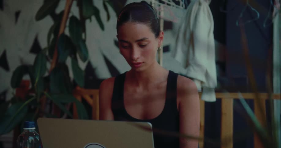 A focused young woman with her hair tied back works intently on her laptop in a dimly lit, stylish cafe