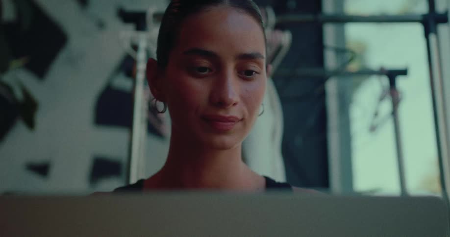 A young woman with her hair pulled back is focused on her laptop in a dimly lit room