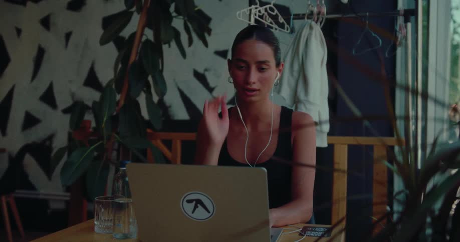 A young woman with earphones participates in a video call on her laptop while working remotely in a stylish cafe