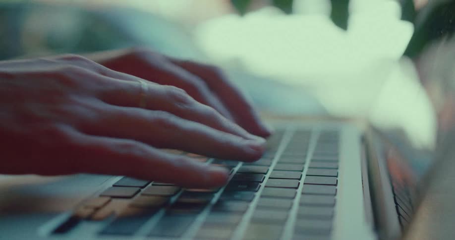 A close-up side view of a person's hands with a wedding ring typing on a laptop keyboard in a softly lit room