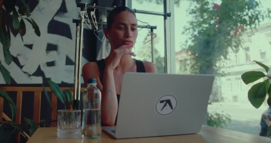 A young woman with earphones thoughtfully works on her laptop in a modern cafe by a large window