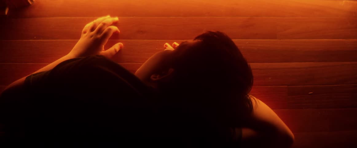 A silhouetted person lies on a wooden floor, dramatically illuminated by a mysterious orange light seeping from under a closed door