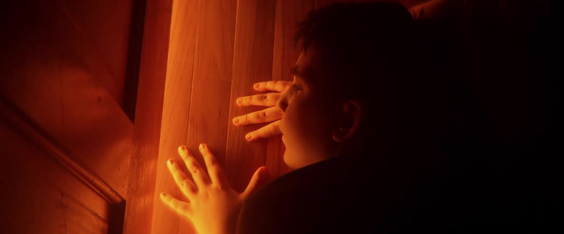 A terrified young boy presses his hands against a wooden door, trapped and hiding in a dark room illuminated by an ominous orange glow