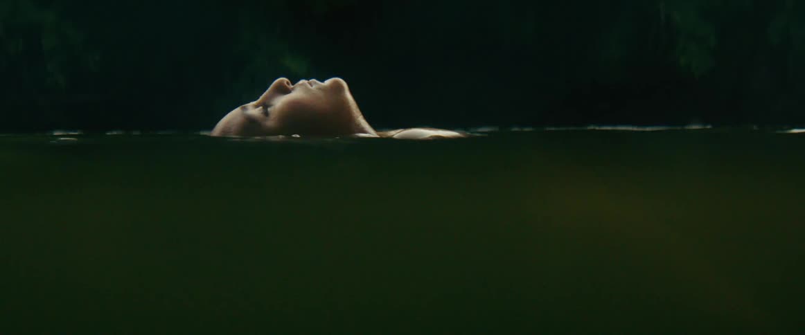 A serene boys face emerges from the dark, calm water of a secluded lake as he floats on her back