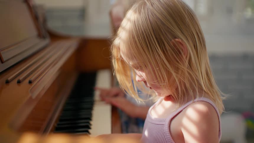 A close-up of two young blonde sisters learning to play the piano together in a sunlit room
