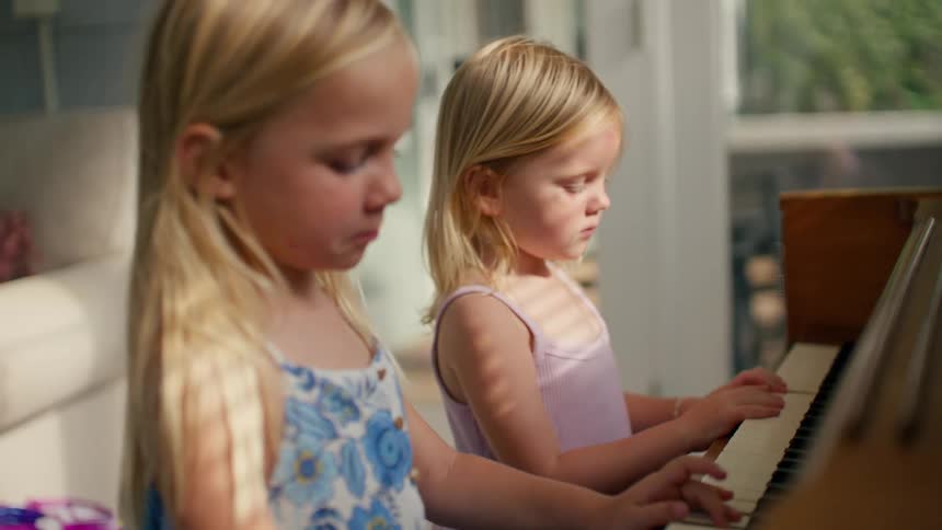 Two young sisters concentrate while learning to play the piano together in a sunlit room