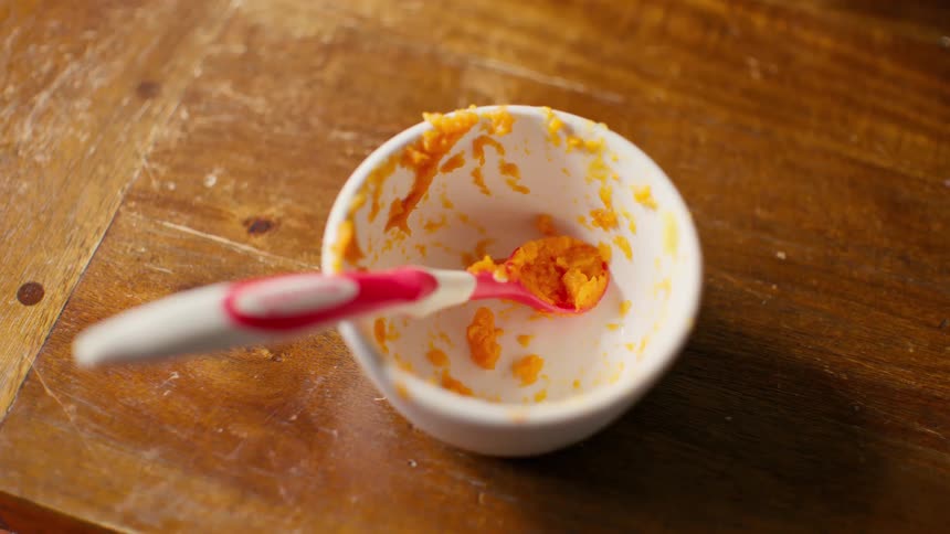 A messy, finished bowl of orange baby food with a spoon sits on a rustic wooden table