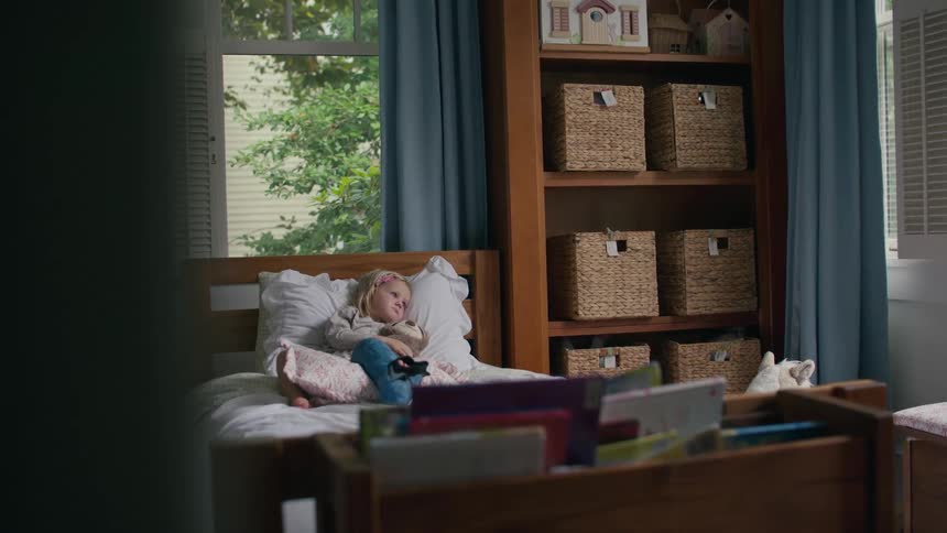 A wide shot shows a little girl resting in her bed, watching a show in her cozy bedroom