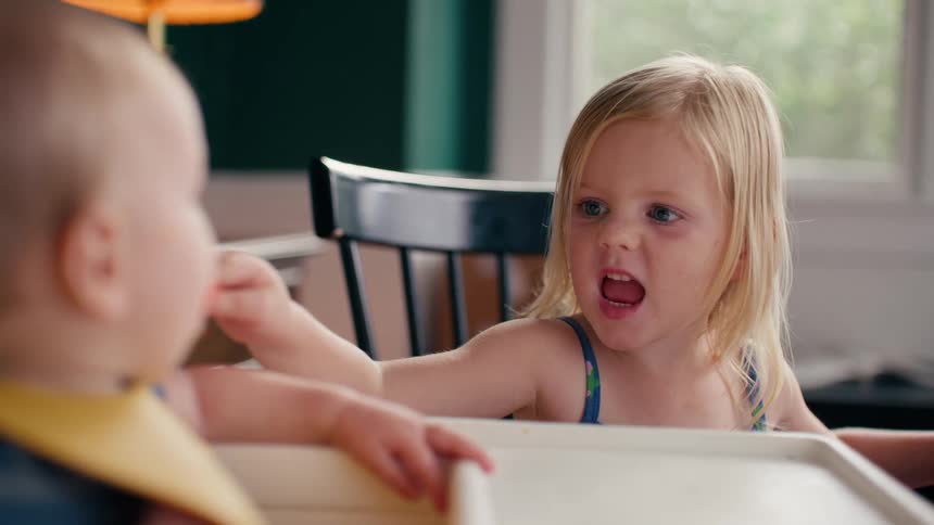 A cute toddler playfully scoops food from a bowl and feeds her baby sibling in a high chair, making a funny roaring face