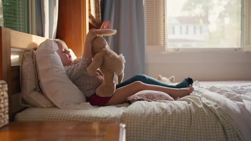 A young girl playfully interacts with her stuffed bunny rabbit while relaxing in her sunlit bed