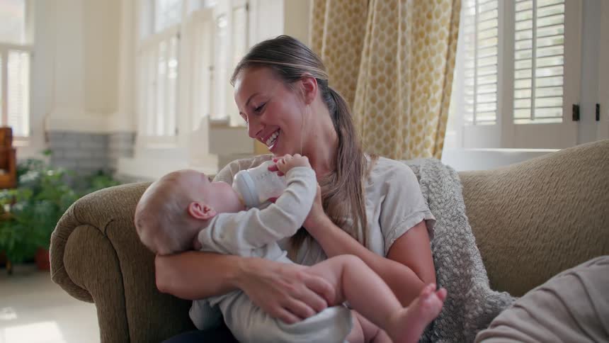 A loving mother smiles as she bottle-feeds her baby while relaxing on the couch in their sunlit living room