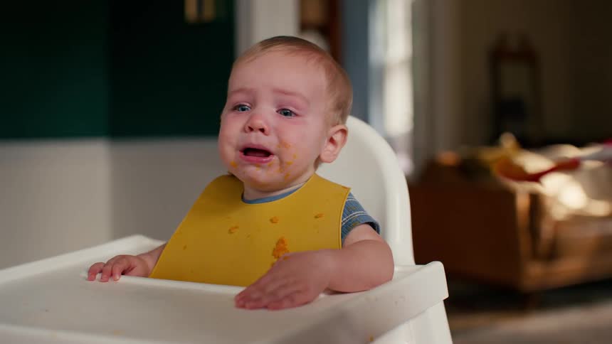An upset baby with a messy face crying while being spoon-fed in a high chair