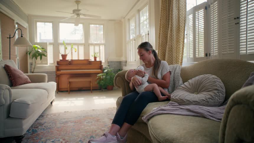 A caring mother bottle-feeds her newborn baby as they relax on the couch in a bright, cozy living room