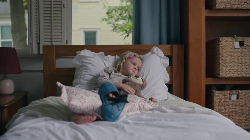A sad little girl with a cast on her leg rests in bed, propped up on pillows with her favorite stuffed animal
