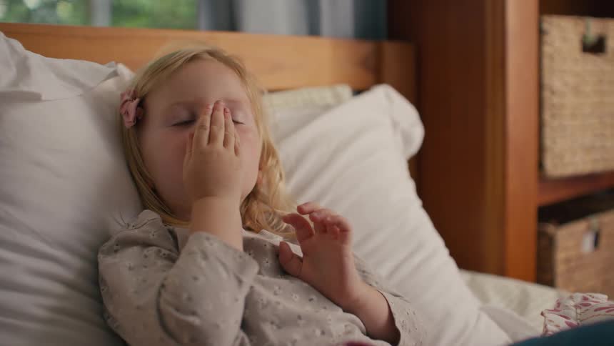 A sleepy little blonde girl rubs her eyes and then smiles as she wakes up from a nap in her bed in slow motion