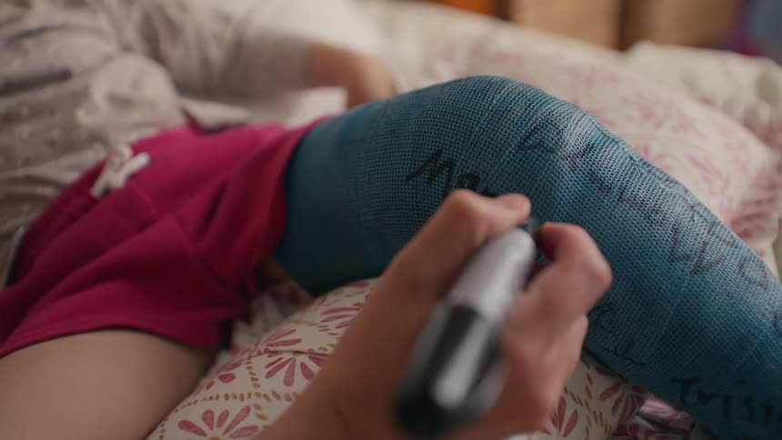 A mother tenderly writes a message on her young daughter's blue leg cast as she recovers from an injury