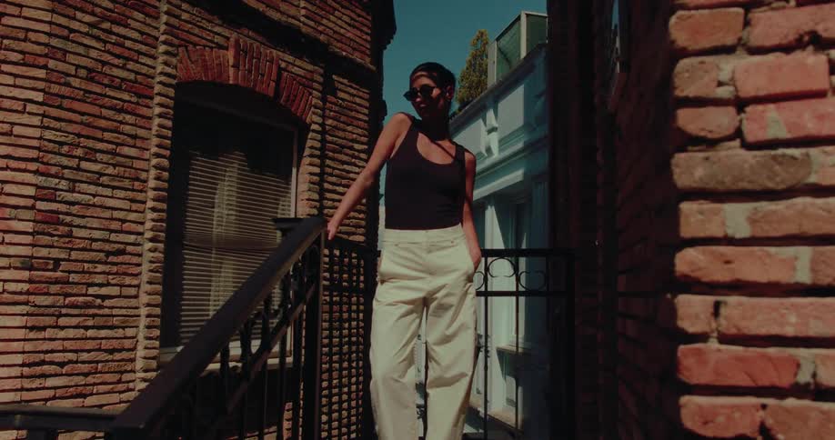 A stylish woman in sunglasses and a black tank top poses confidently on a balcony in a narrow brick alleyway on a sunny day