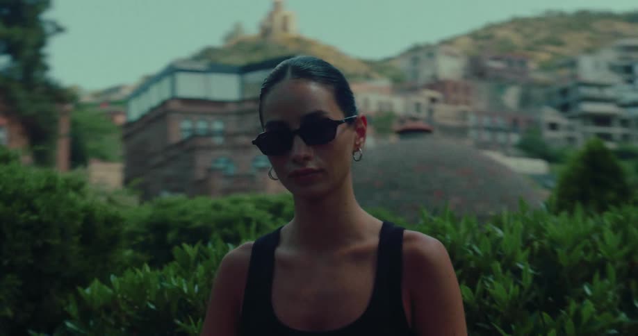 A stylish woman in sunglasses looks into the camera as it slowly reveals the historic domed buildings of an old city in the background