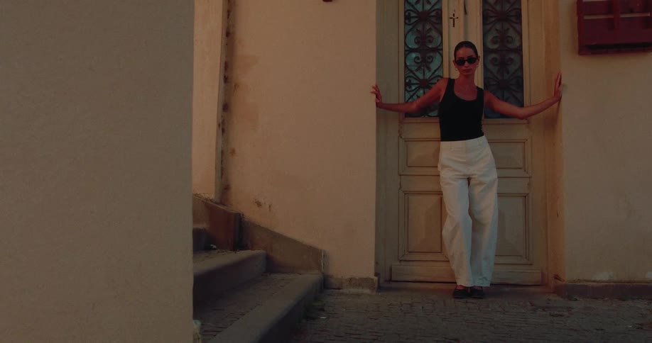 A fashionable woman in sunglasses and white pants poses dramatically against an old door in a narrow street at dusk