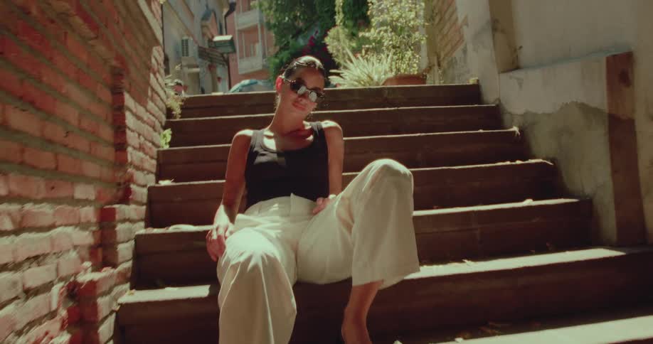 A fashionable woman in sunglasses poses while sitting on wooden stairs in a sunny European alleyway