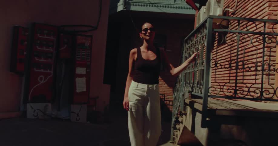 A stylish woman in sunglasses poses in the sunlight of a shadowy European-style courtyard