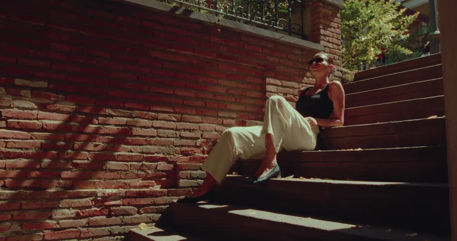 A fashionable woman in sunglasses relaxes on a sunlit staircase next to a brick wall