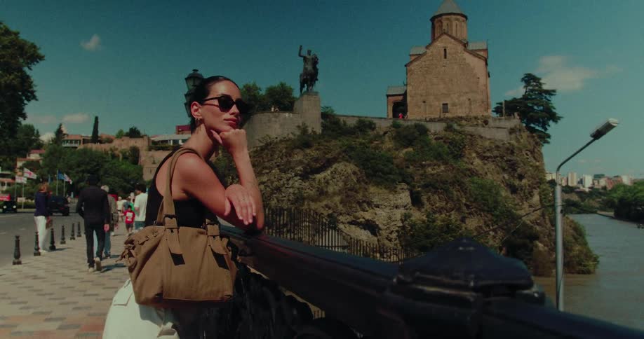 A stylish woman in sunglasses stands on a bridge, thoughtfully taking in the historic view of Metekhi Church in Tbilisi, Georgia