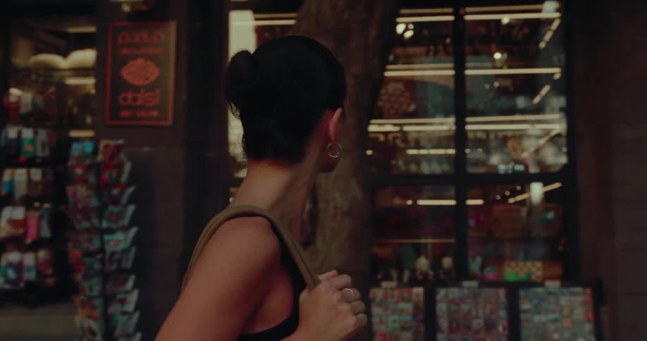 A beautiful young woman with dark hair in a bun walks down a city street past brightly lit shops in slow motion