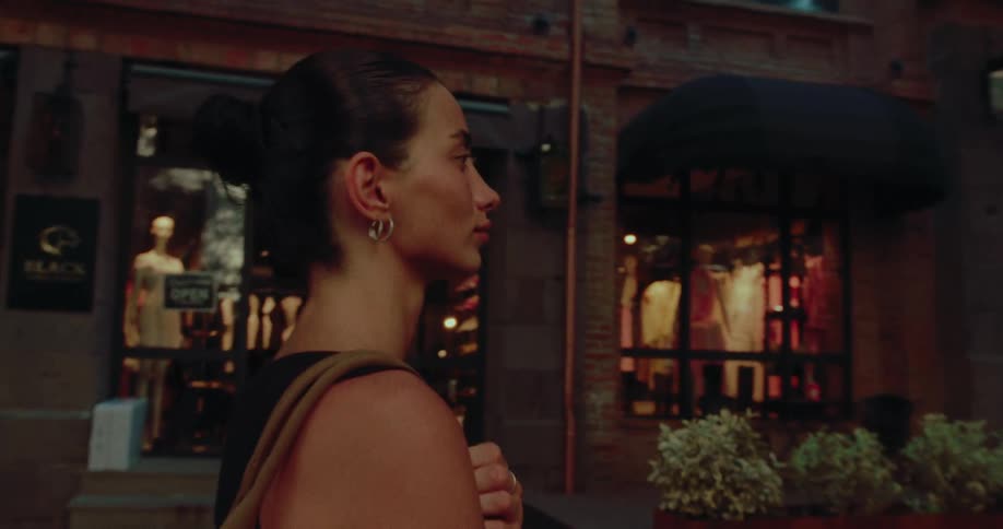 A stylish woman walks down an urban street in the evening, passing the warm glow of a boutique storefront